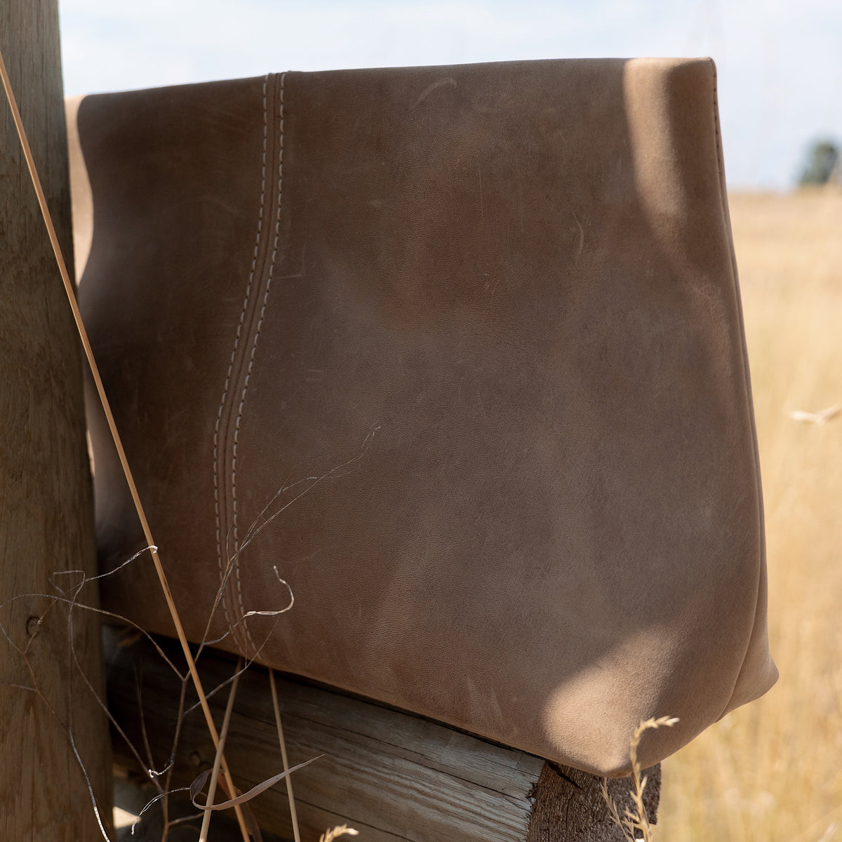A brown leather bag with visible stitching, leaning against a rustic wooden fence post in an outdoor field under a cloudy sky.